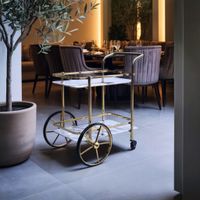 Luxury serving trolley with champagne gold brushed stainless steel frame, white sintered stone shelves, and black wheels, styled in a dining area with wine bottles, glassware, and a green indoor plant