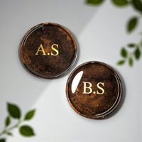 A pair or resin coasters, brown in colour with gold monogram initials. resting on a white background
