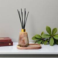 A resin reed diffuser with black reeds resting on a matching oval dish, they are both beige tones in colour. Both resting on a white shelf with a plant
