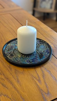 round resin display plate with a peacock style design and black edging. There is a candle resting on top
