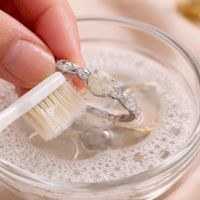 A close-up of a person's fingers holding a silver ring with a light-colored center stone over a glass bowl of soapy water, gently scrubbing it with a white toothbrush.